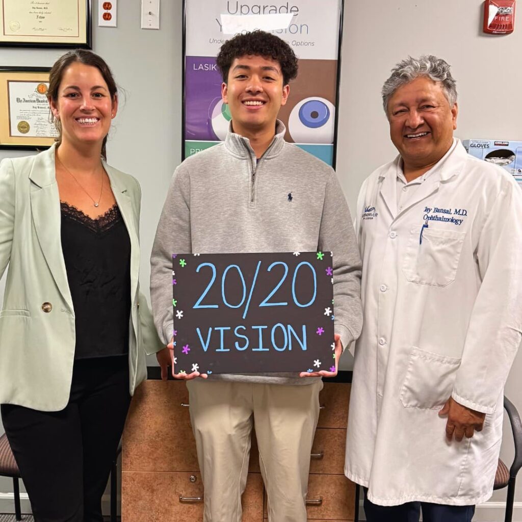 Happy SMILE post-op patient at LaserVue Eye Center stands with Dr. Jay Bansal and Dr. Francesca Sefton, holding a sign that reads "20/20 Vision"