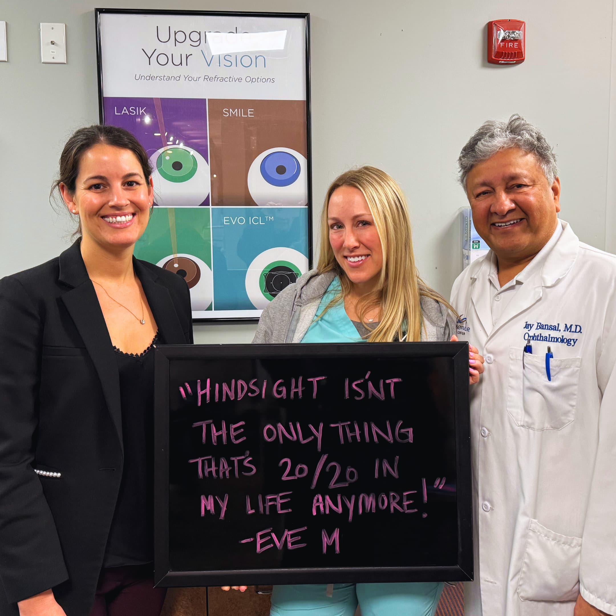 Eve M. LASIK Patient at LaserVue Eye Center standing with Dr. Jay Bansal and Dr. Francesca Sefton, holding a sign that reads "Hindsight isn't the only thing that's 20/20 in my life anymore! Eve M."