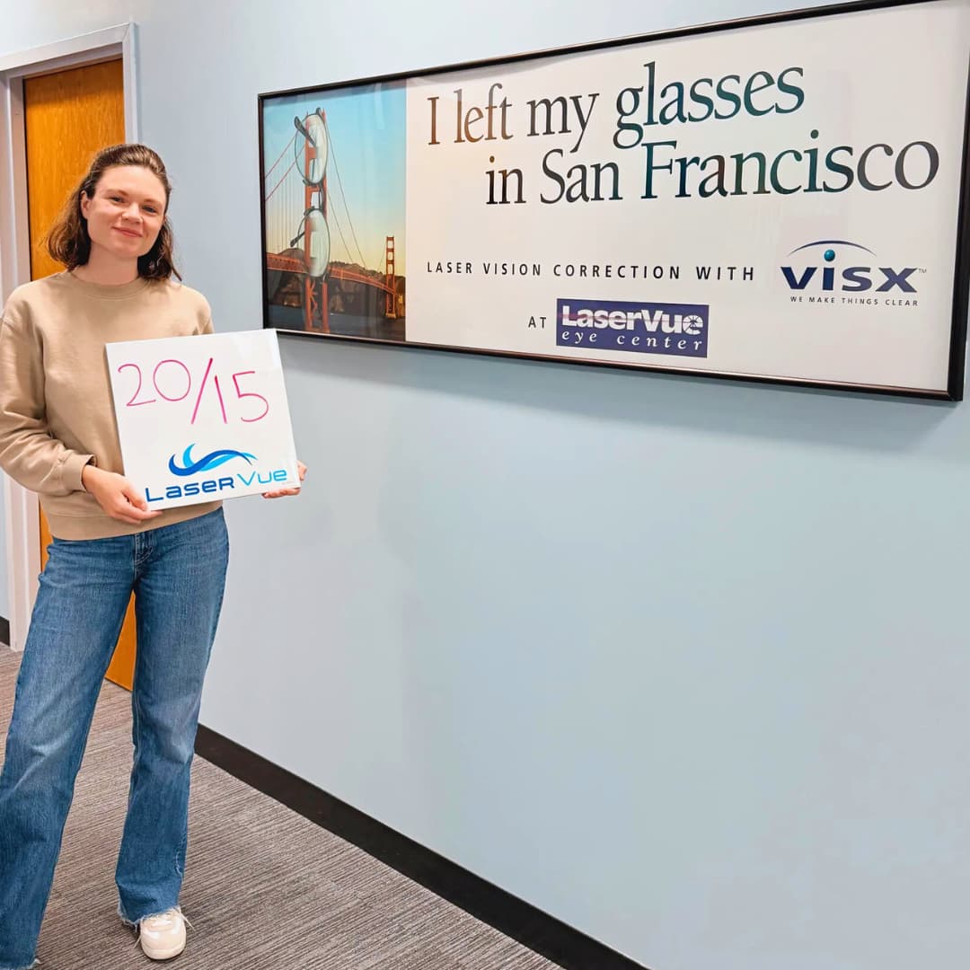 20/15 vision post-LASIK patient from LaserVue Eye Center holds a sign while standing in front of another sign. The sign in her hands reads "20/15. LaserVue." The sign behind her reads "I left my glasses in San Francisco. Laser Vision Correction with LaserVue Eye Center"