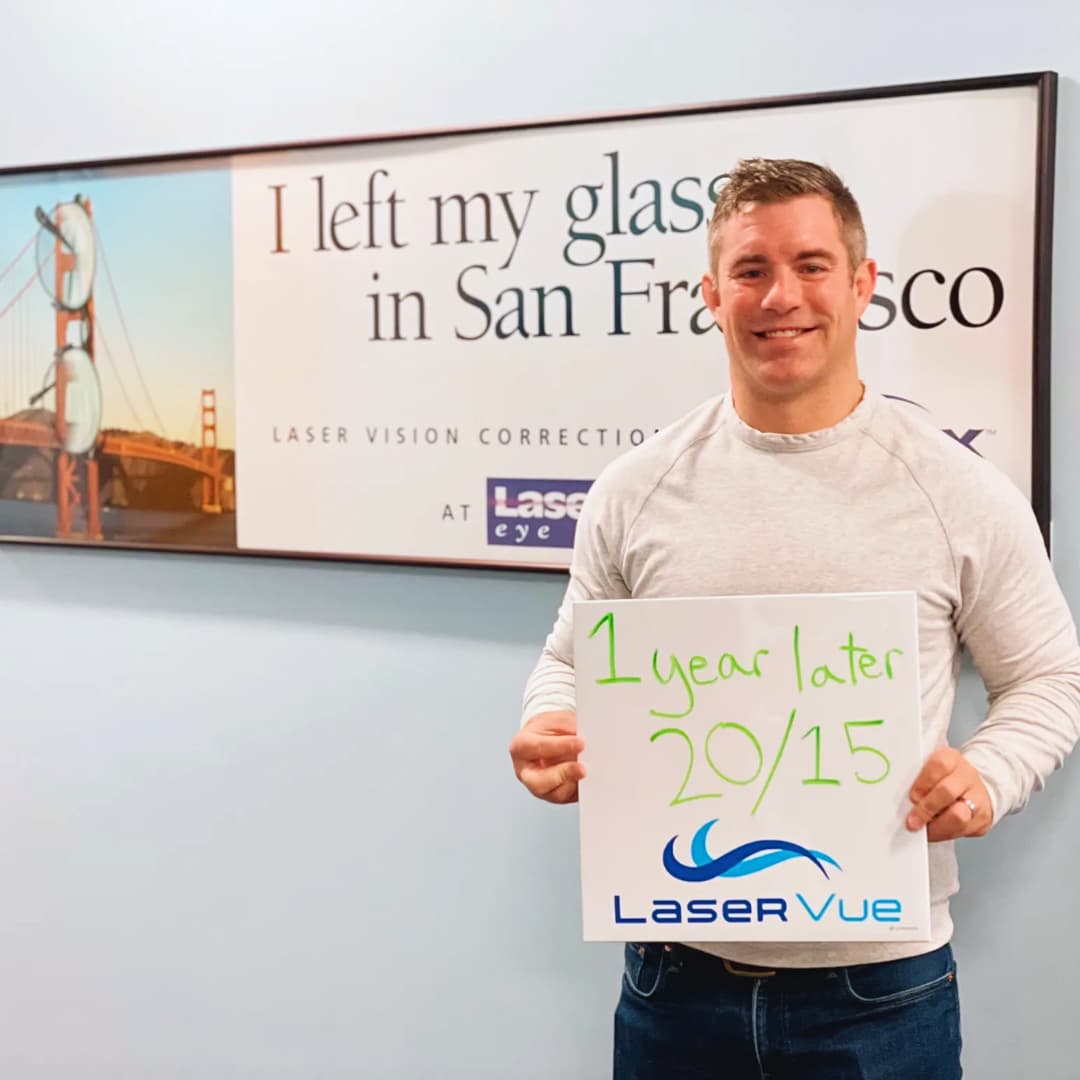 Happy LASIK patient stands in front of a sign, holding a whiteboard. The sign reads "I left my glasses in San Francisco. Laser Vision Correction with LaserVue Eye Center". The whiteboard reads "1 year later. 20/15. LaserVue"