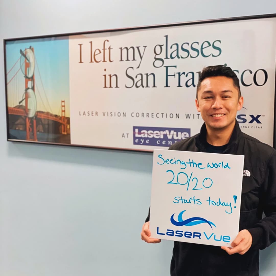 Happy man stands in front of a sign that reads "I left my glasses in San Francisco. Laser Vision Correction with LaserVue Eye Center". He is holding a white board that reads "Seeing the world 20/20 starts today! LaserVue"