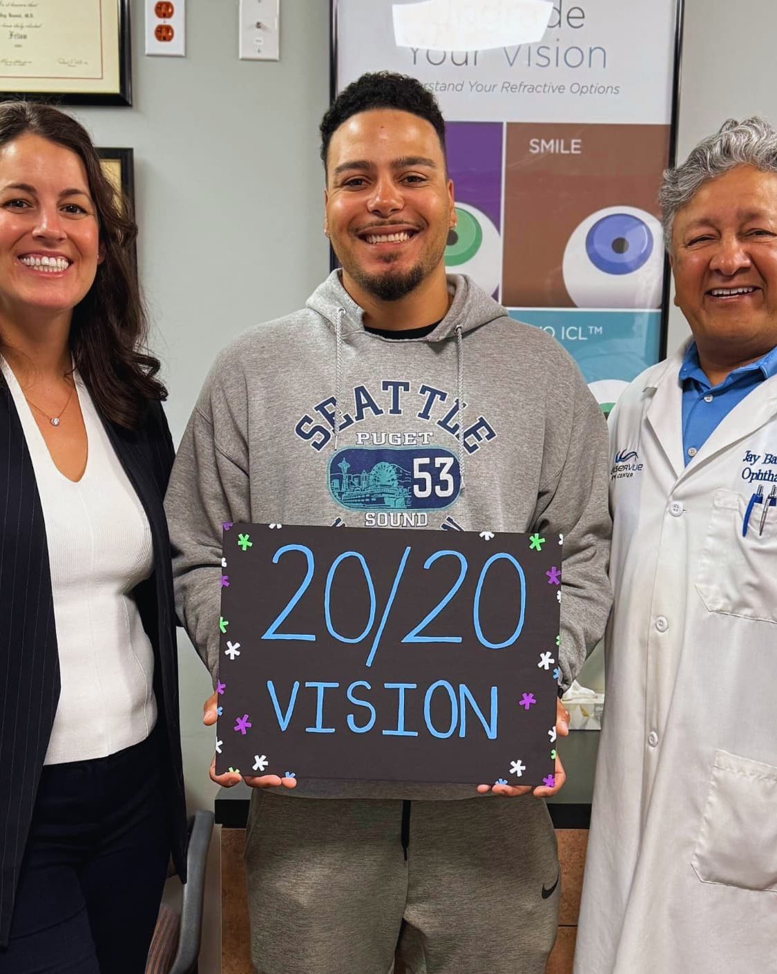 Happy LASIK post-op patient stands next to Dr. Jay Bansal and Dr. Francesca Sefton. The patient is holding a sign that reads "20/20 vision".