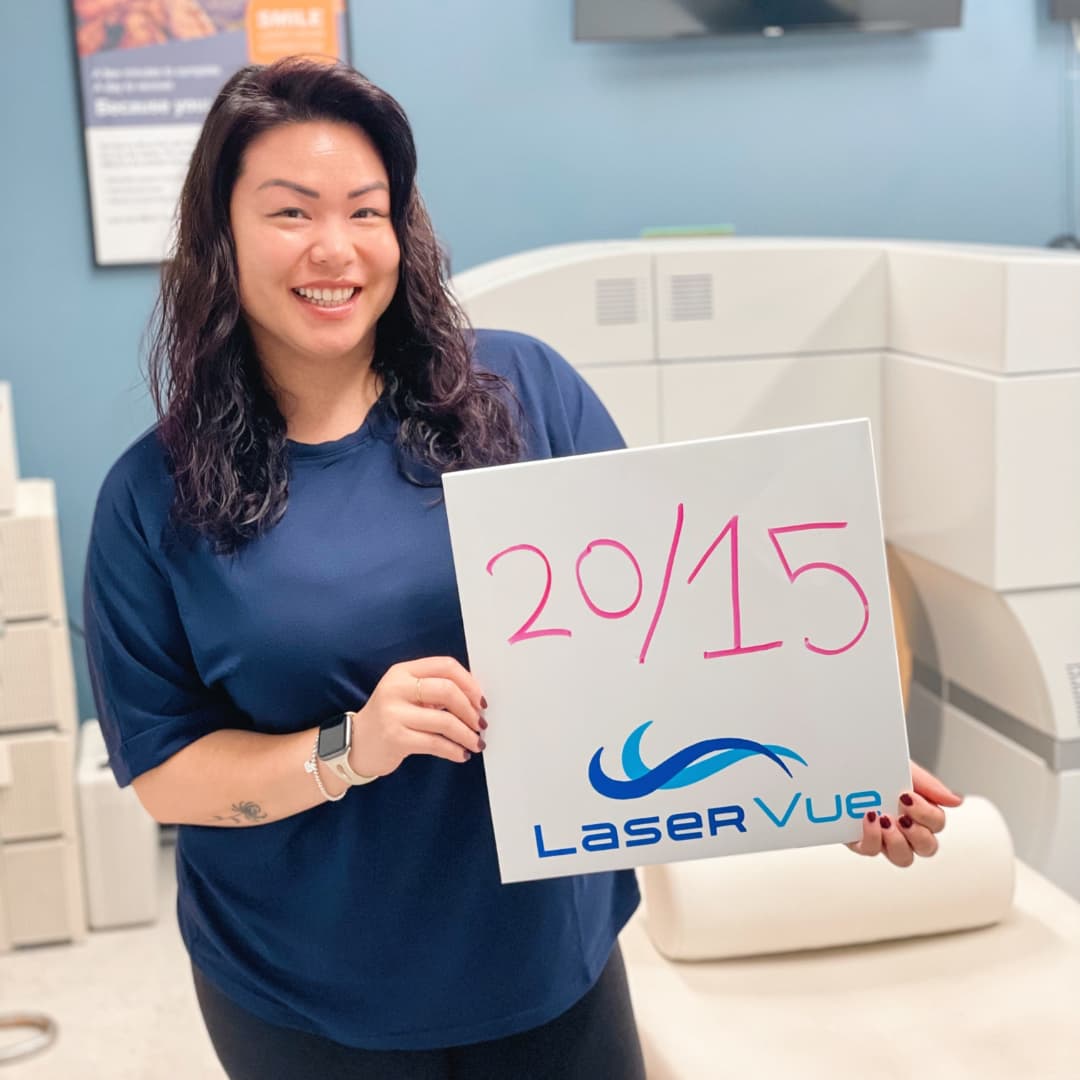 Happy woman stands in front of a ZEISS SMILE femtosecond laser, holding a sign that reads "20/15. LaserVue"
