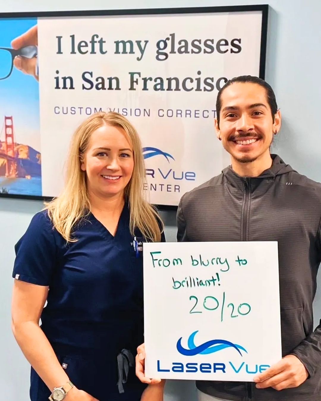 LASIK patient at LaserVue Eye Center in San Francisco smiles while standing with Dr. Hannah Walsh, optometrist. The patient holds a sign that reads "From blurry to brilliant! 20/20. LaserVue." The patient and optometrist are standing in front of a poster that reads "I left my glasses in San Francisco. Custom Vision Correction with LaserVue Eye Center."