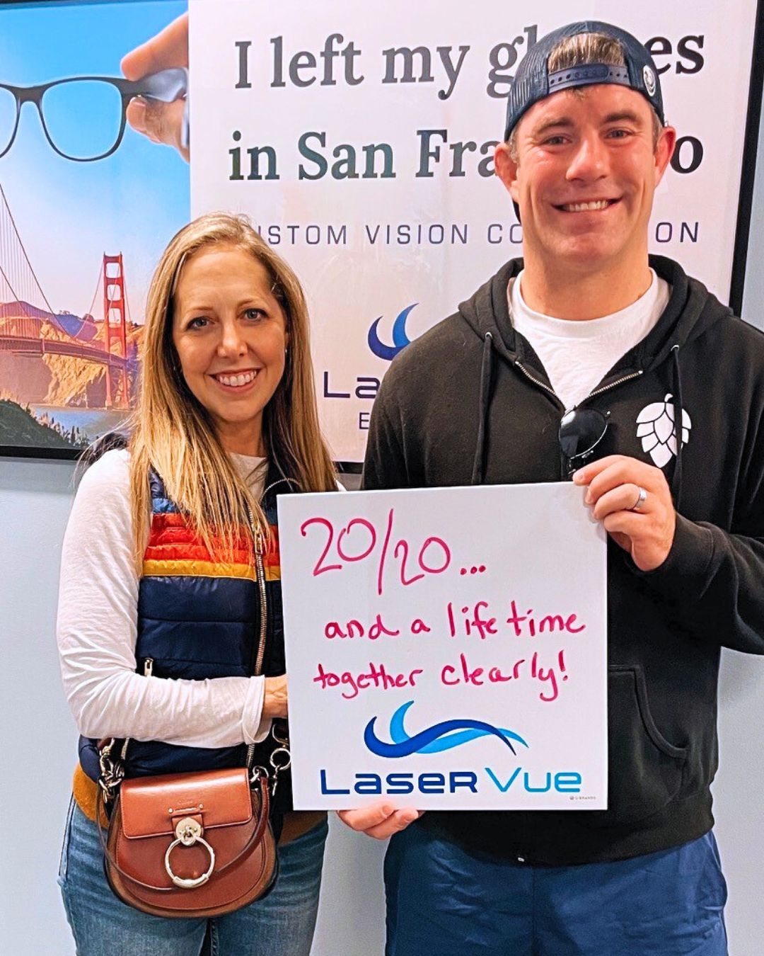 Happy LASIK patient, Laura W. celebrates her LASIK surgery and 20/20 vision at LaserVue Eye Center. She is with her husband, Scott L. who also had LASIK at LaserVue. She holds a whiteboard that reads "20/20... And a lifetime together clearly! LaserVue." Behind the couple is a poster that reads "I left my glasses in San Francisco. Custom vision correction with LaserVue Eye Center."