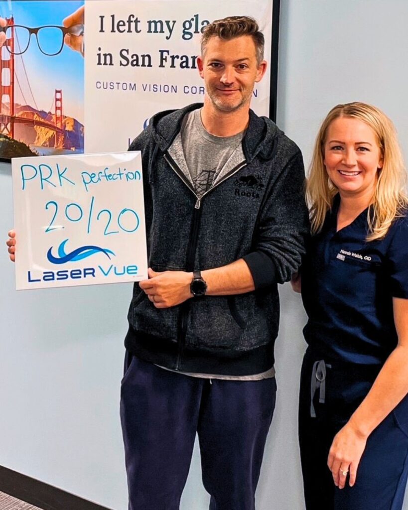 PRK patient at LaserVue Eye Center in San Francisco smiles while standing with Dr. Hannah Walsh, optometrist. The patient holds a sign that reads "PRK Perfection. 20/20. LaserVue." The patient and optometrist are standing in front of a poster that reads "I left my glasses in San Francisco. Custom Vision Correction with LaserVue Eye Center."