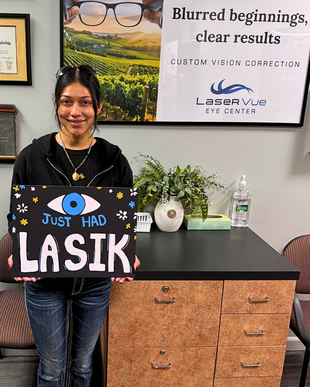 Happy LaserVue LASIK patient, Maria S., stands in the reception of LaserVue Eye Center in Santa Rosa. She is holding a sign that reads "Just Had LASIK" with a drawing of an eye and fireworks. Poster on the wall reads "Blurred beginnings, clear results. Custom vision correction with LaserVue Eye Center."