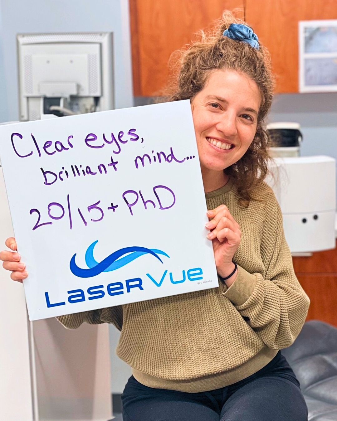Happy woman sits during a post-operative exam for LASIK surgery at LaserVue Eye Center. She holds a sign that reads "Clear eyes, brilliant mind... 20/15 + PhD. LaserVue."