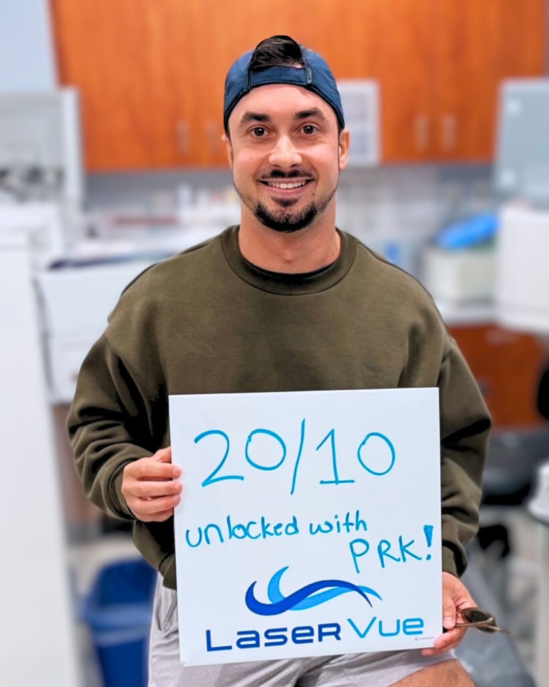 Happy LaserVue PRK Patient smiles at his post-op appointment. He is holding a sign that reads "20/10 unlocked with PRK! LaserVue."