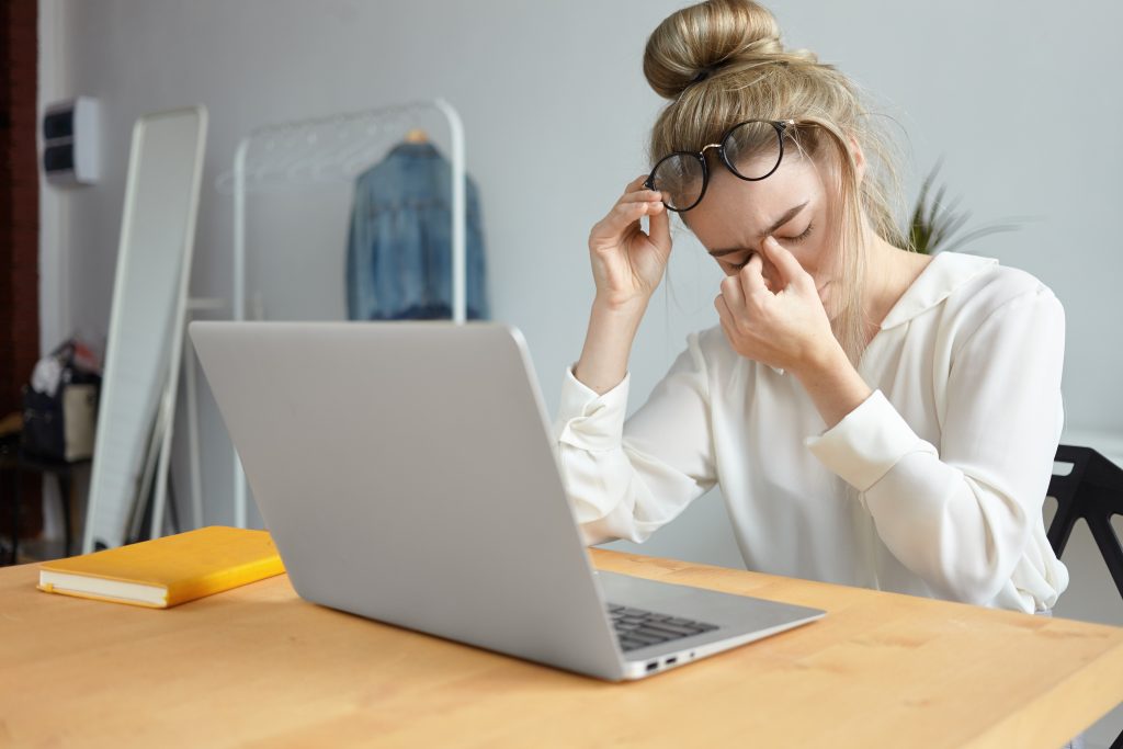 Woman at a laptop removing her glasses and rubbing her eyes from strain