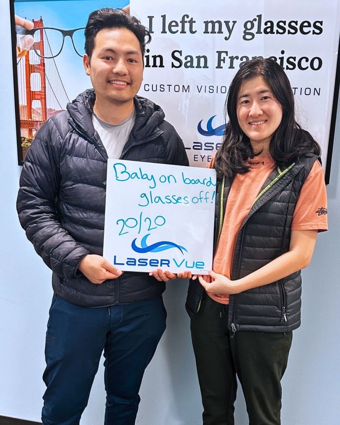 Travis V. and Sophia Z., married couple and LASIK patients at LaserVue Eye Center in San Francisco. Smiling while Holding a sign that reads "Baby on board, glasses off! 20/20. LaserVue."