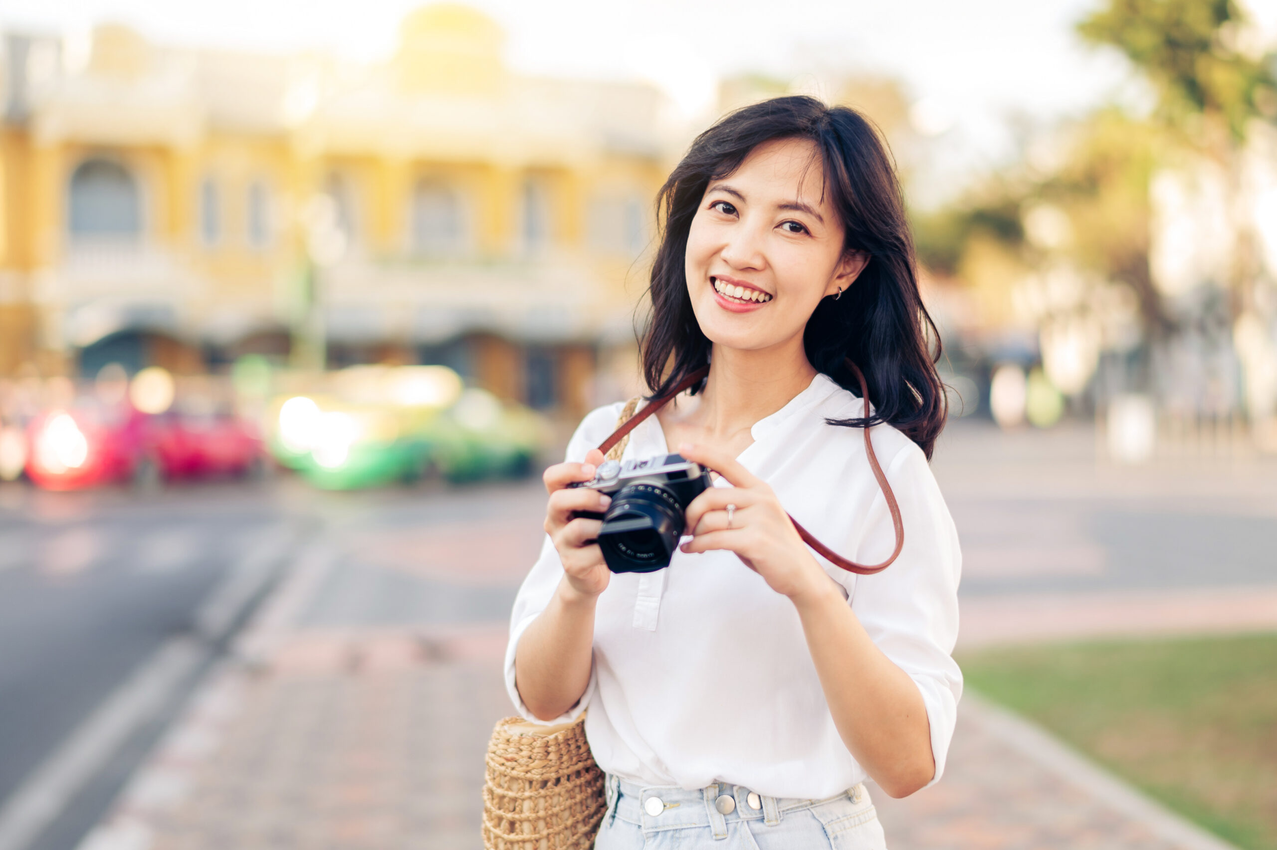 Woman smiles at camera holding a photography camera, while on vacation in the daytime. She is wearing a white shirt and jeans.