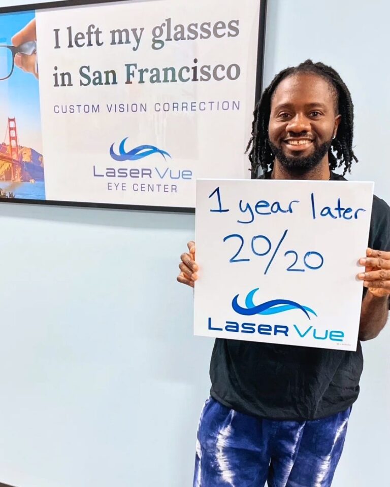 PRK patient at LaserVue Eye Center, Adolphus B., attends his 1-year post op exam. He stands smiling while holding a whiteboard that reads "1 year later 20/20. LaserVue." He stands in front of a poster that reads "I left my glasses in San Francisco. Custom vision correction with LaserVue Eye Center."
