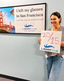 20/15 vision post-LASIK patient Alison S. from LaserVue Eye Center holds a sign while standing in front of another sign. The sign in her hands reads "20/15. LaserVue." The sign behind her reads "I left my glasses in San Francisco. Laser Vision Correction with LaserVue Eye Center"
