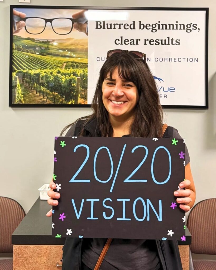 LASIK Patient at LaserVue Eye Center, Angelina F., holds a sign that reads "20/20 vision". Behind her is a poster reading "Blurred beginnings, clear results. Custom vision correction with LaserVue Eye Center."