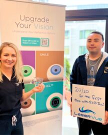 Happy EVO ICL patient Christian R. celebrates his 20/20 vision at LaserVue Eye Center. Dr. Hannah Walsh, optometrist is standing next to him holding a model of the EVO implantable collamer lens. The man holds a whiteboard that reads "20/20 for the first time with EVO ICL! LaserVue." Behind them is a poster that reads "Upgrade Your Vision: Understanding Your Refractive Options. LASIK. SMILE. PRK. EVO ICL."