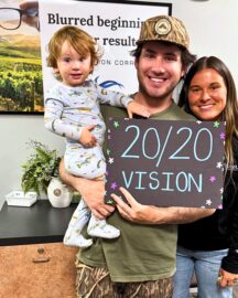 Happy LASIK patient, Christopher B., stands with his partner and child, holding a hand painted sign that reads "20/20 vision." The poster behind him reads "Blurred beginnings, clear results. Custom vision correction with LaserVue Eye Center."