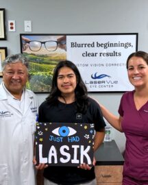 Happy LaserVue LASIK patient, Jesus R., stands with ophthalmologist and LASIK surgeon Dr. Jay Bansal and optometrist Dr. Francesca Sefton. Jesus holds a sign that reads "Just Had LASIK" with a drawing of an eye and fireworks. They all stand in front of a poster that reads "Blurred beginnings, clear results. Custom vision correction with LaserVue Eye Center."