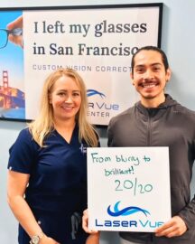 LASIK patient at LaserVue Eye Center in San Francisco smiles while standing with Dr. Hannah Walsh, optometrist. The patient holds a sign that reads "From blurry to brilliant! 20/20. LaserVue." The patient and optometrist are standing in front of a poster that reads "I left my glasses in San Francisco. Custom Vision Correction with LaserVue Eye Center."