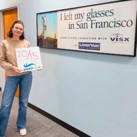 20/15 vision post-LASIK patient from LaserVue Eye Center holds a sign while standing in front of another sign. The sign in her hands reads "20/15. LaserVue." The sign behind her reads "I left my glasses in San Francisco. Laser Vision Correction with LaserVue Eye Center"