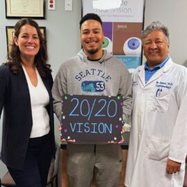 Happy LASIK post-op patient stands next to Dr. Jay Bansal and Dr. Francesca Sefton. The patient is holding a sign that reads "20/20 vision".