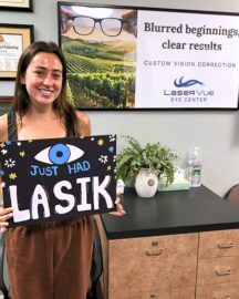 Happy LASIK Patient from LaserVue Eye Center stands with a blackboard that says "(Eye) Just Had LASIK". She is standing in front of a poster that reads "Blurred beginnings, clear results. Custom Vision Correction. LaserVue Eye Center."