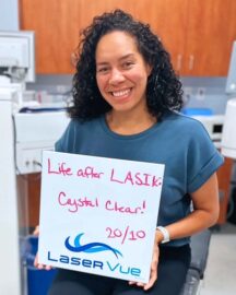LASIK post-op patient at LaserVue Eye Center, Sheridan N., sits in the surgery room where she received LASIK. She is holding a sign that reads "Life after LASIK crystal clear! 20/10. LaserVue."