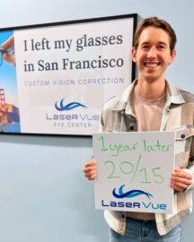 Happy LASIK patient attends his 1-year post-op exam at LaserVue Eye Center. He smiles while holding a whiteboard that reads "1 year later. 20/15. LaserVue." Behind him is a poster that reads "I left my glasses in San Francisco. Custom vision correction with LaserVue Eye Center."