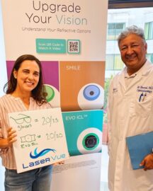 Happy EVO ICL patient Trish S. celebrates her 20/20 vision at LaserVue Eye Center. Dr. Jay Bansal, EVO ICL surgeon and ophthalmologist is standing next to her. Trish holds a whiteboard that reads "Glasses = 20/25. EVO ICL = 20/20. LaserVue." Behind them is a poster that reads "Upgrade Your Vision: Understanding Your Refractive Options. LASIK. SMILE. PRK. EVO ICL."